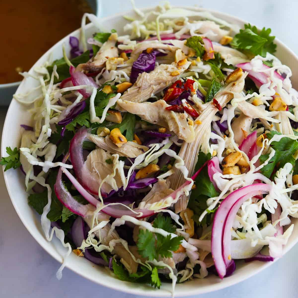 Image of cabbage salad with chicken and garnished with onions, cilantro, mint, peanuts.