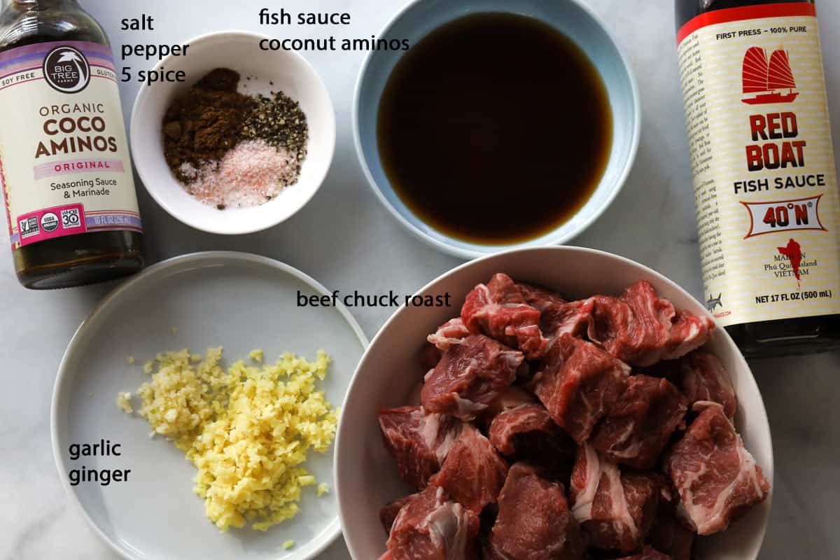 Picture of ingredients. Coconut aminos bottle, spices, fish sauce bottle, minced garlic and ginger and cut beef.