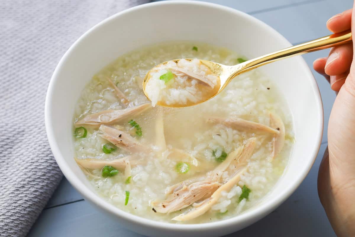 A bowl of rice porridge with shredded chicken, garnished with green onions. Spoon of rice porridge lifting up from bowl.