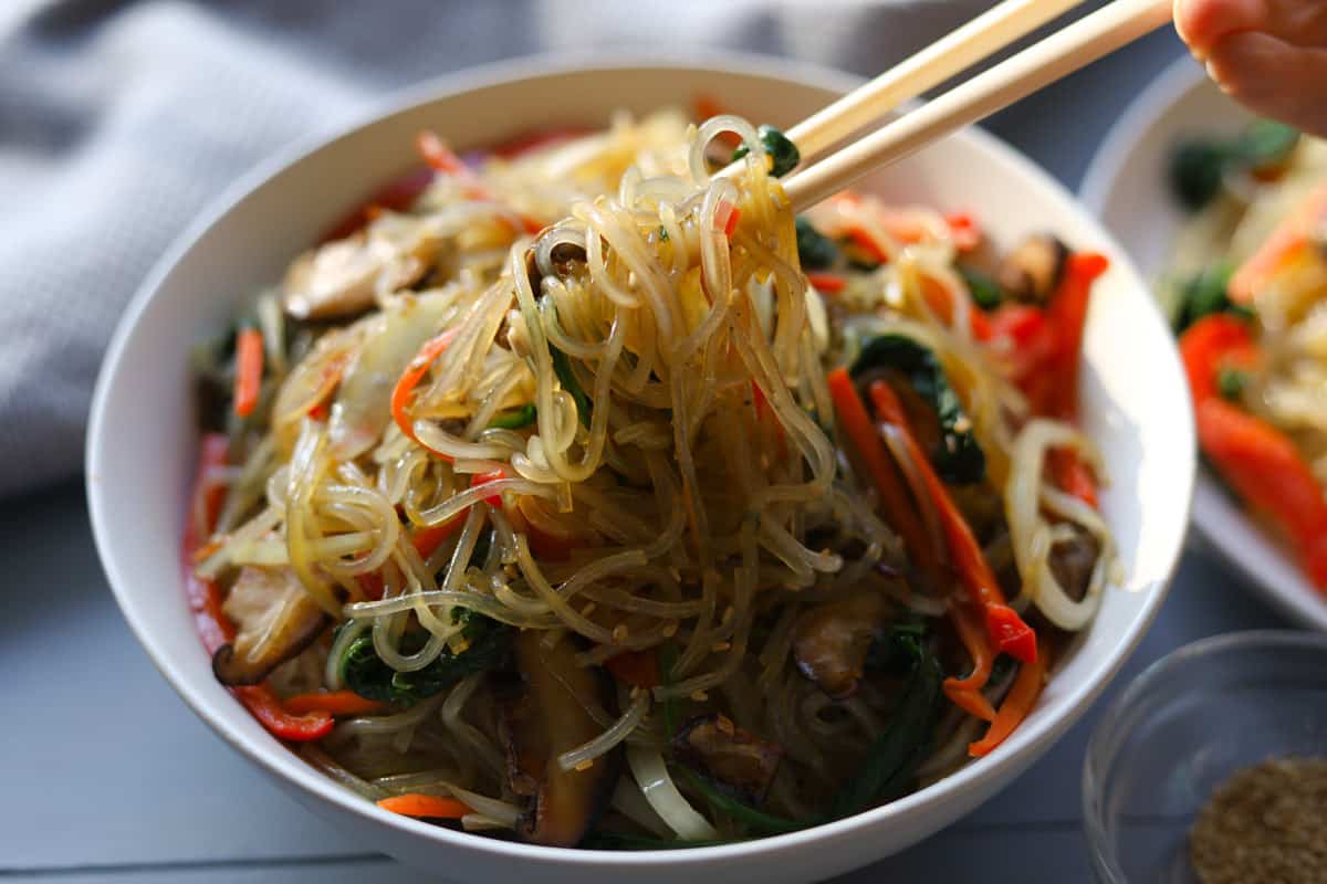 Bowl of sweet potato noodles with mixed vegetables and beef. Chopsticks lifting noodles out of bowl.  