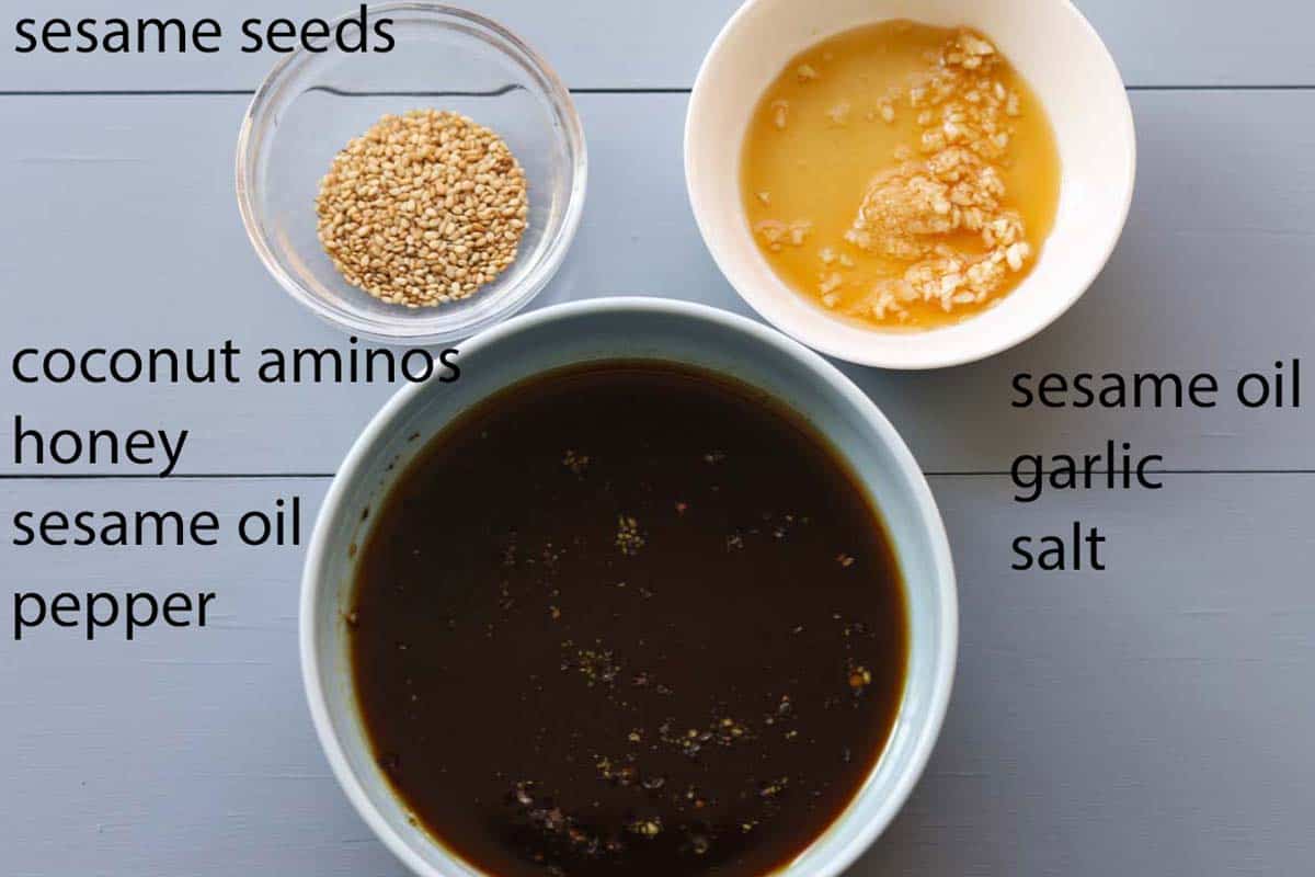 Image of ingredients. Sesame seeds in small bowl, sesame oil, garlic and salt in small bowl. Coconut aminos mixed with honey, sesame oil and pepper in small bowl. 