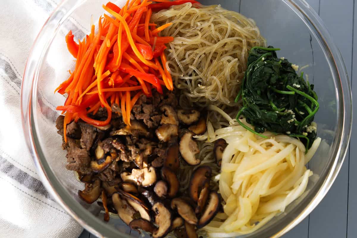 Image of sweet potato noodles, cooked carrots, bell peppers, spinach, mushrooms, beef and onions in a bowl. 
