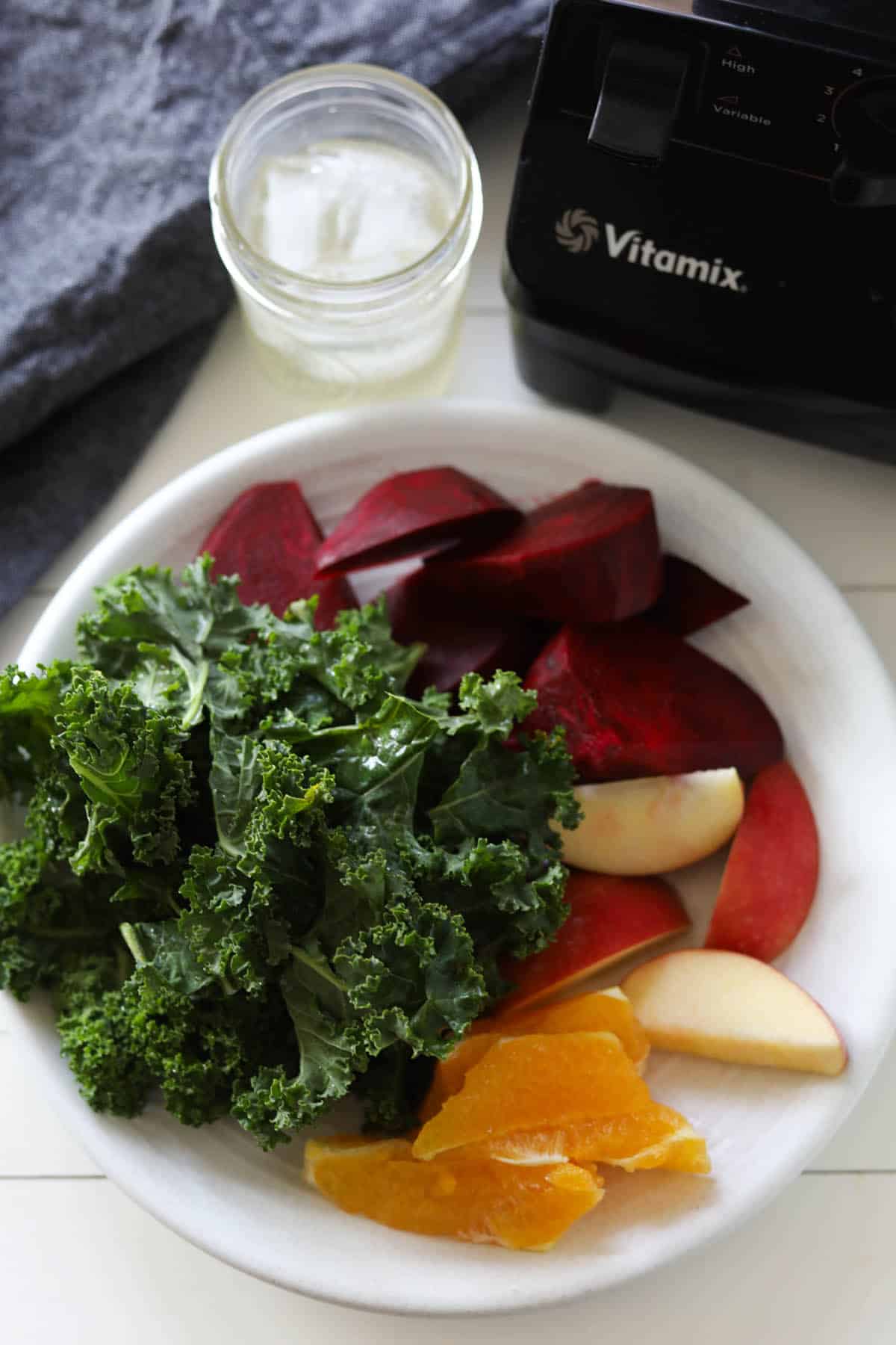 Image of blender in background and cup of ice and clear liquid. Plate of chopped green kale, cut red beet, apple and orange slices.