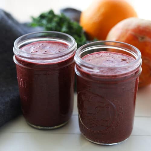 Image of two small glass cups filled with red smoothie. Fruit and kale in background.