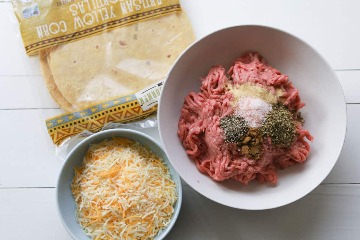 Ingredients on a table. Tortillas, cheese in a bowl, ground beef with seasoning in a bowl.