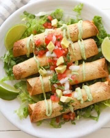 Four golden taquitos on top a bed of sliced lettuce, drizzled with avocado crema sauce and topped with diced tomatoes, cilantro, onions and avocado.