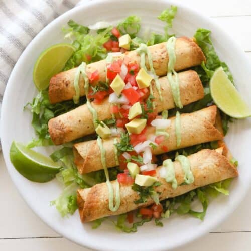 Four golden taquitos on top a bed of sliced lettuce, drizzled with avocado crema sauce and topped with diced tomatoes, cilantro, onions and avocado.