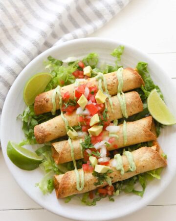 Four golden brown taquitos on a bed of sliced lettuce, garnished with avocado crema, diced tomatoes, onions, avocado and cilantro.