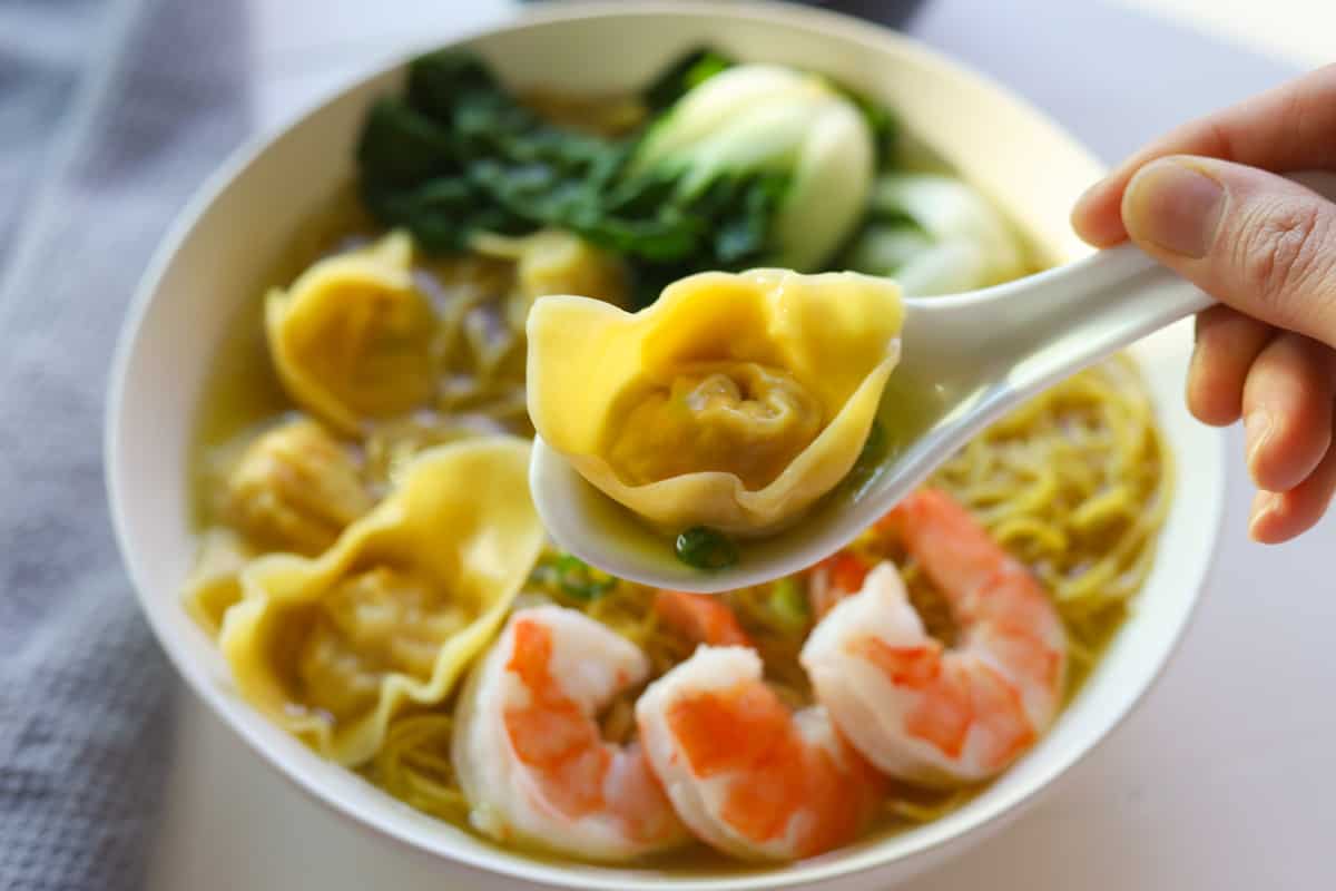 Closeup of spoon holding a single wonton. Background of bowl of wonton soup with noodles, shrimp, bokchoy and green onions.