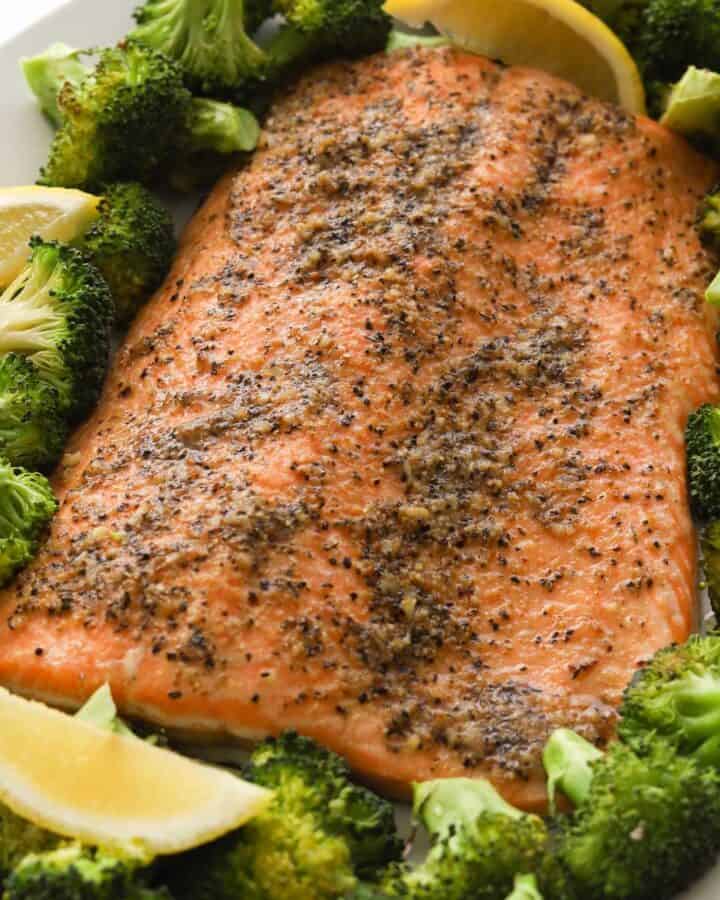Closeup image of cooked salmon fillet with broccoli florets and lemon wedges on a plate.