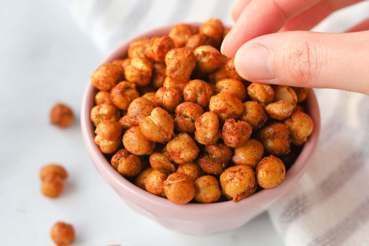 Close up image of fingers picking up roasted chickpeas with seasoning from a bowl.