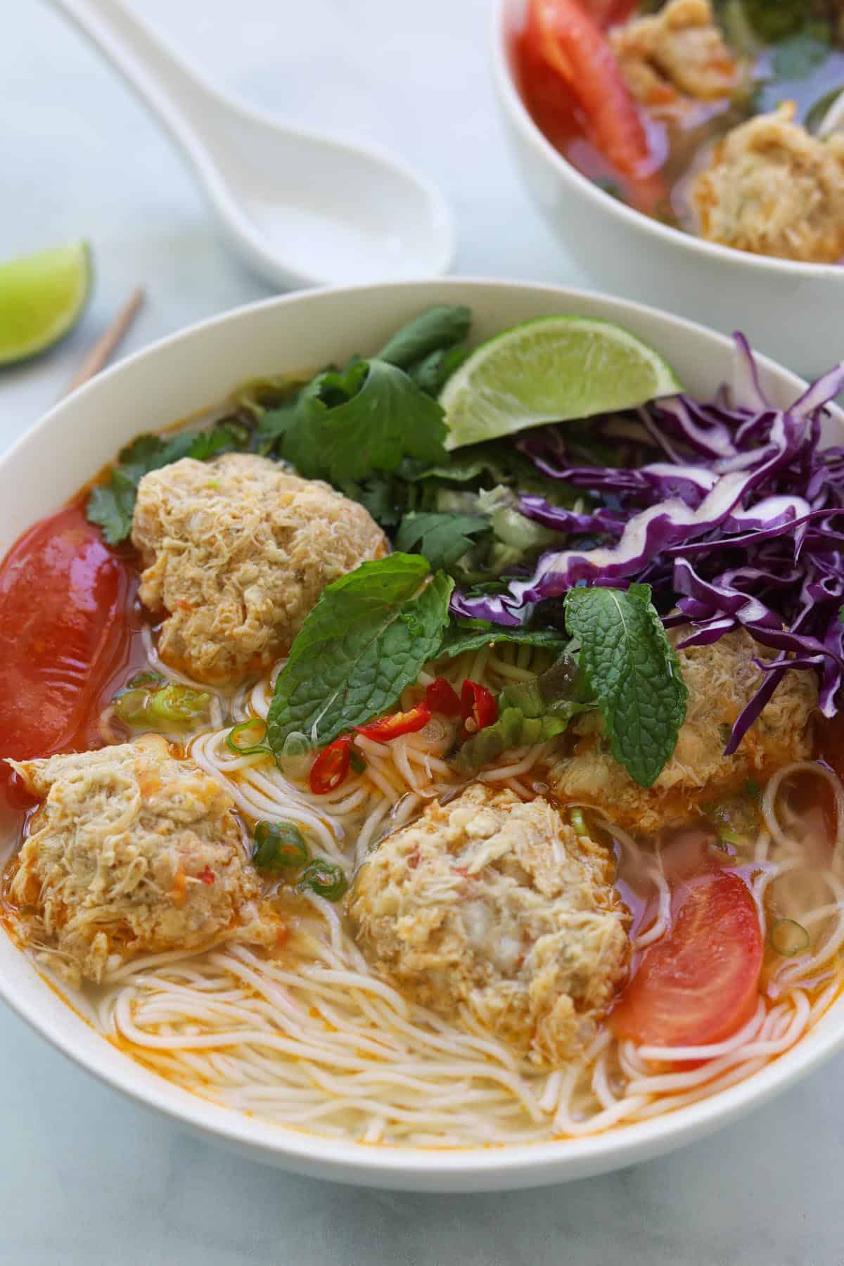Bowl of rice noodles with soup, crab meatballs, tomatoes and garnished with fresh greens and herbs.