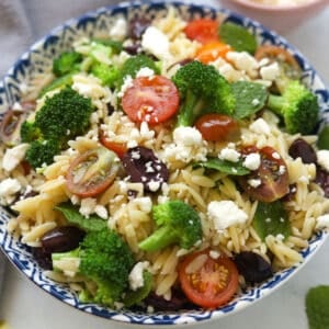 Bowl of orzo pasta salad with broccoli, tomatoes, mint leaves, olives and feta cheese.