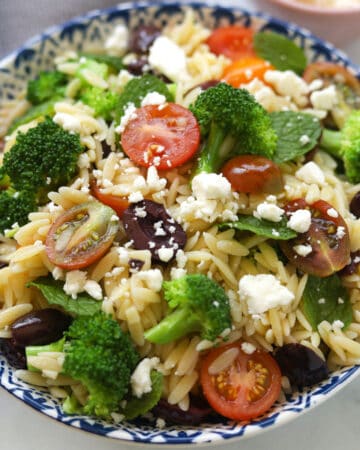 Bowl of orzo pasta salad with broccoli, tomatoes, mint leaves, olives and feta cheese.