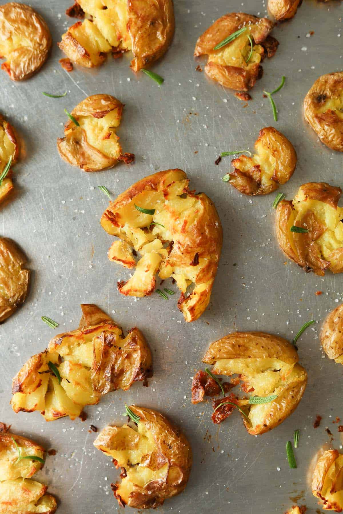 Roasted smashed potatoes with rosemary leaves on roasting pan.