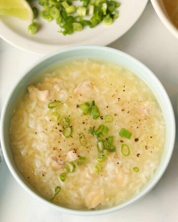 Bowl chicken rice porridge with sliced scallions and sliced limes on side.