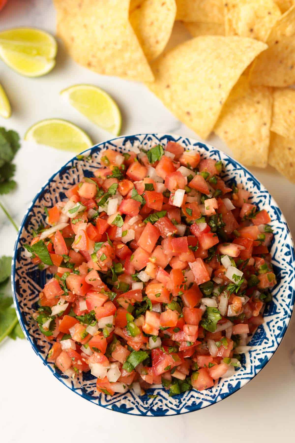 pico de gallo salsa in bowl with sliced limes, tortilla chips and cilantro in background.