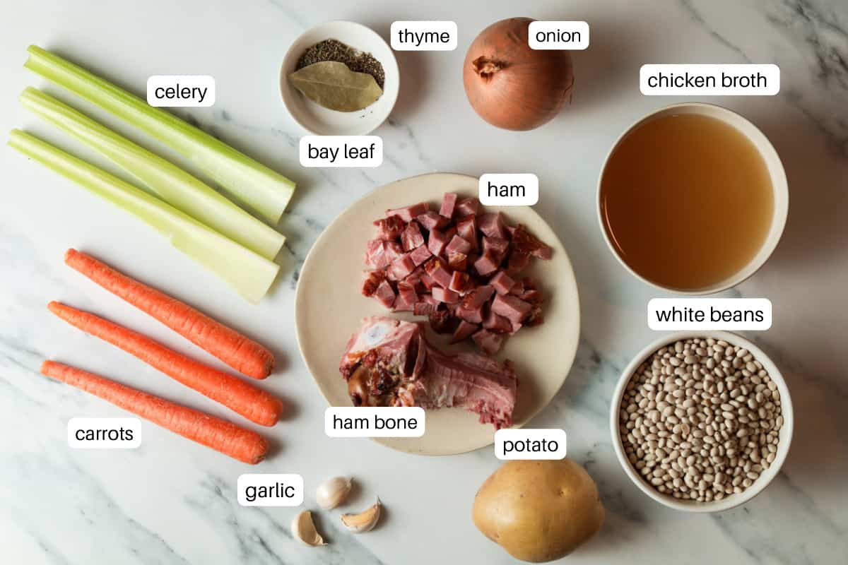 Ingredients on table; carrots, garlic, ham bone, potato, white beans, chicken broth, onion, ham, onion, thyme, bay leaf, celery.