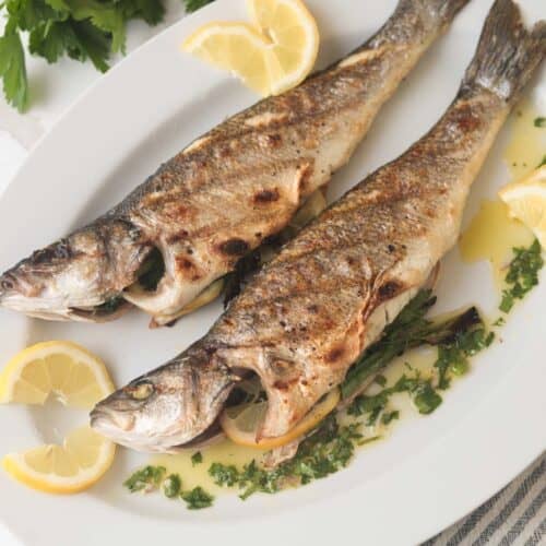 Two whole grilled branzino on plate with herb sauce.
