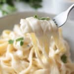 Creamy fettuccine Alfredo being lifted from a ceramic bowl with a silver fork, garnished with fresh parsley.