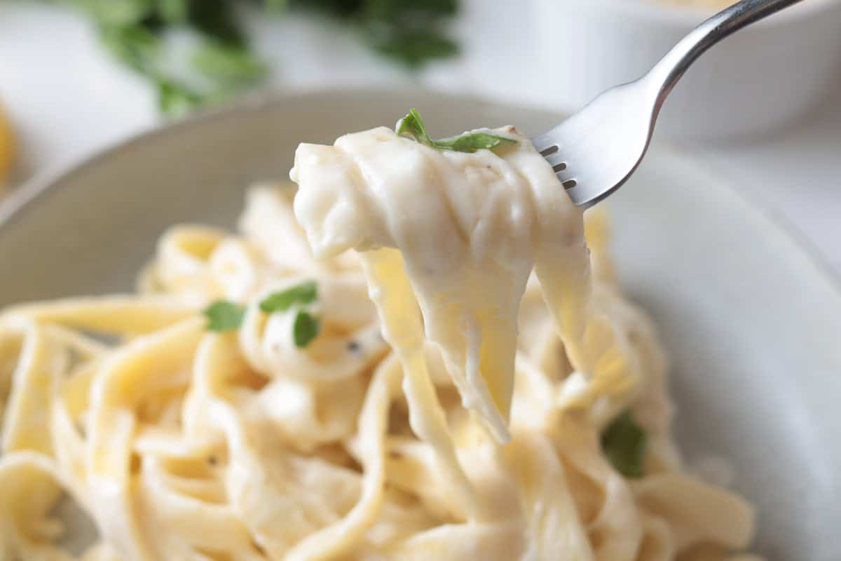 Creamy fettuccine Alfredo being lifted from a ceramic bowl with a silver fork, garnished with fresh parsley.