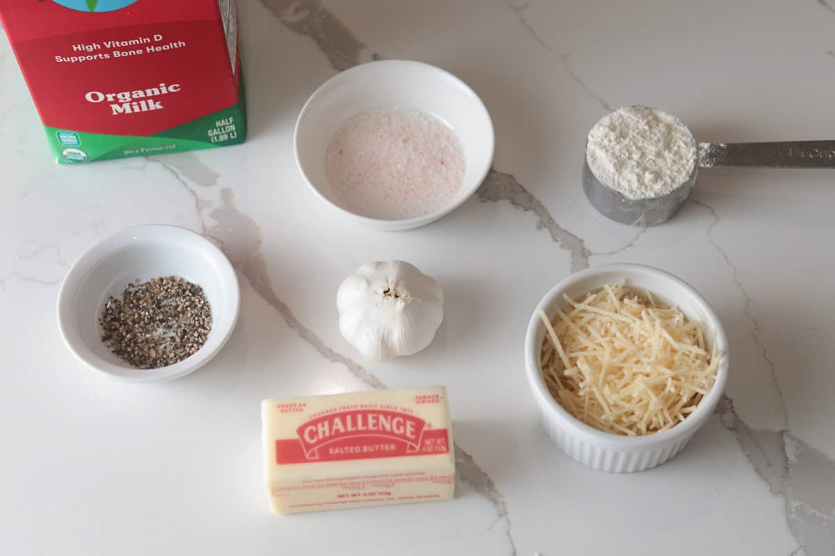 An overhead shot of recipe ingredients, including a carton of organic milk, a stick of butter, garlic, and small bowls of flour, shredded parmesan cheese, salt, and black pepper.