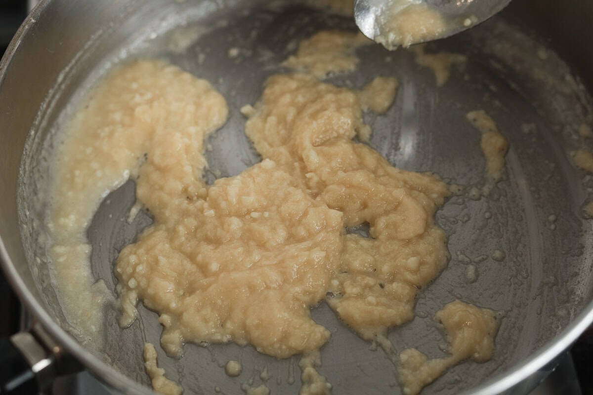 A close-up view looking into a stainless steel pan, showing a thick, pale-golden roux being stirred with a silver spoon.