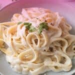 A close-up shot of a plate of pasta tossed in a thick, rich white Alfredo sauce, topped with fresh parsley leaves and ground black pepper. The image has a soft pinkish-purple lighting overlay. Bold white text at the top reads "HOMEMADE ALFREDO SAUCE RECIPE" and "NO HEAVY CREAM NEEDED!", with the website URL www.apeachyplate.com centered at the bottom.