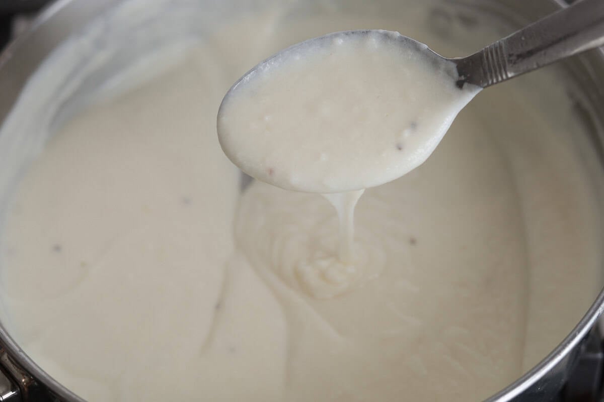 A silver spoon lifting a scoop of smooth, thick white Alfredo sauce from a cooking pot.