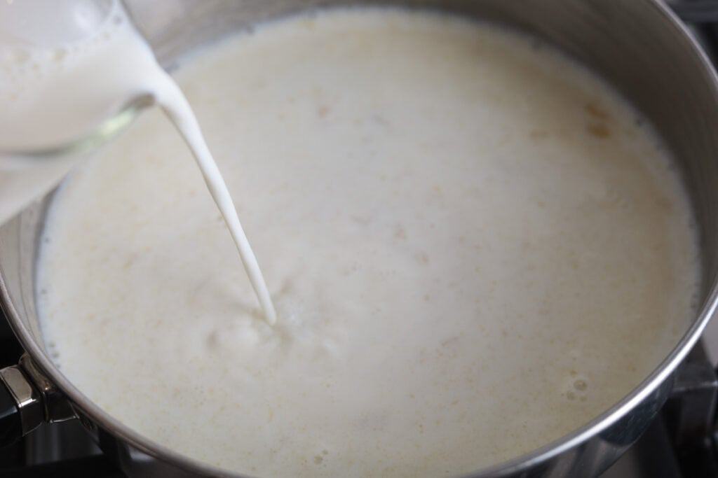 A close-up shot of liquid milk being poured from a glass measuring cup into a stainless steel pan to make Alfredo sauce.