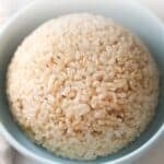 A close-up, high-angle shot of a perfectly shaped dome of fluffy brown rice served in a light blue ceramic bowl. The image text reads "HOW TO COOK THE PERFECT BROWN RICE" at the top and "TRIED & TESTED NO-FAIL METHOD" at the bottom, with the website URL www.apeachyplate.com at the very bottom.