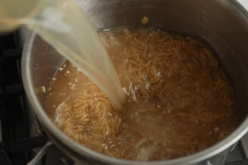 Liquid chicken broth being poured into a pot containing orzo pasta and minced garlic.