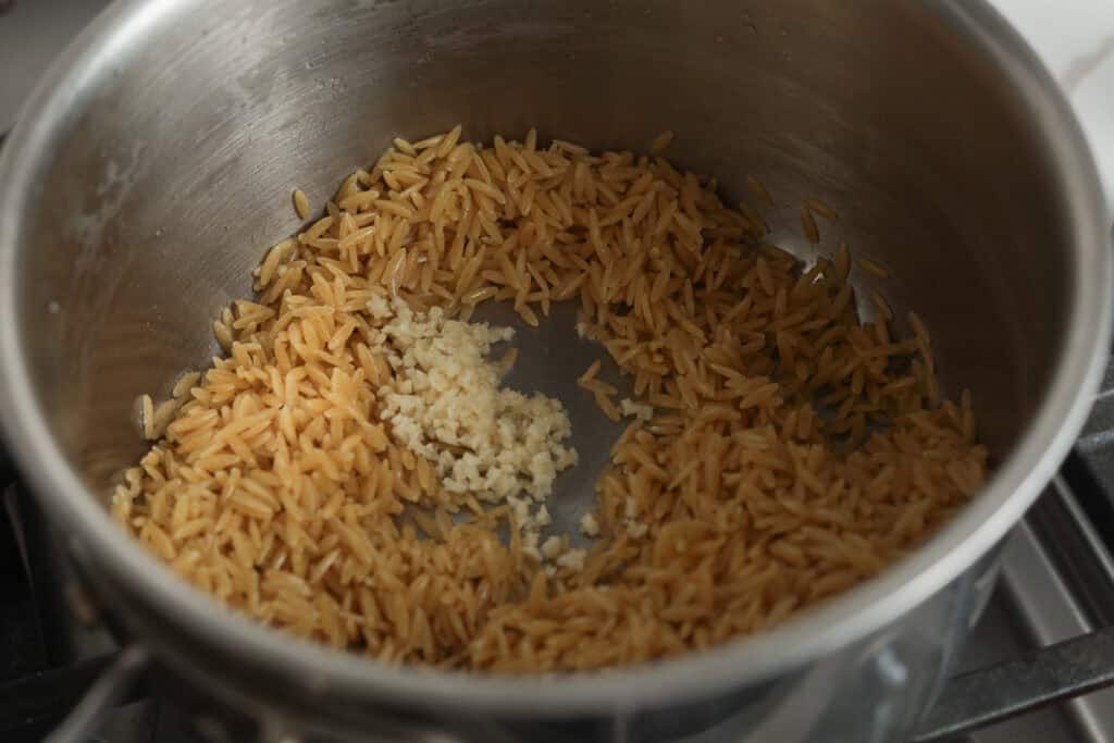 Minced garlic mixed into dry orzo pasta in a pot.