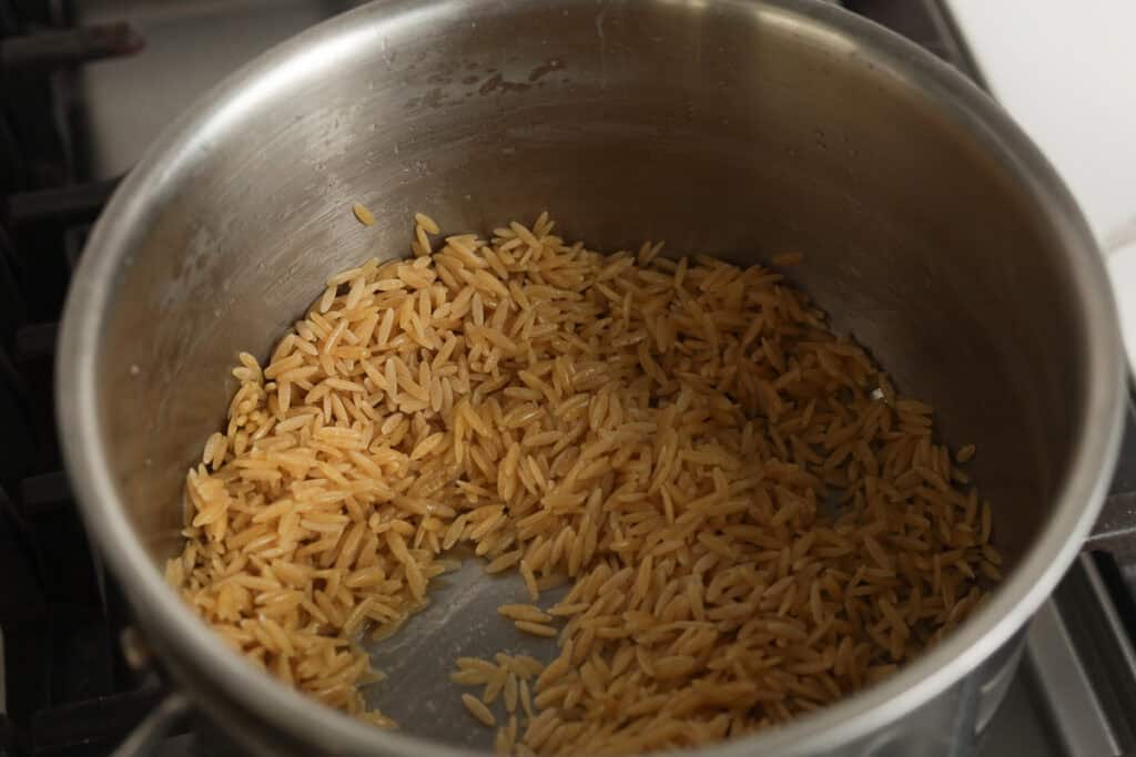 Uncooked dry orzo pasta sitting in a stainless steel saucepan over medium heat.