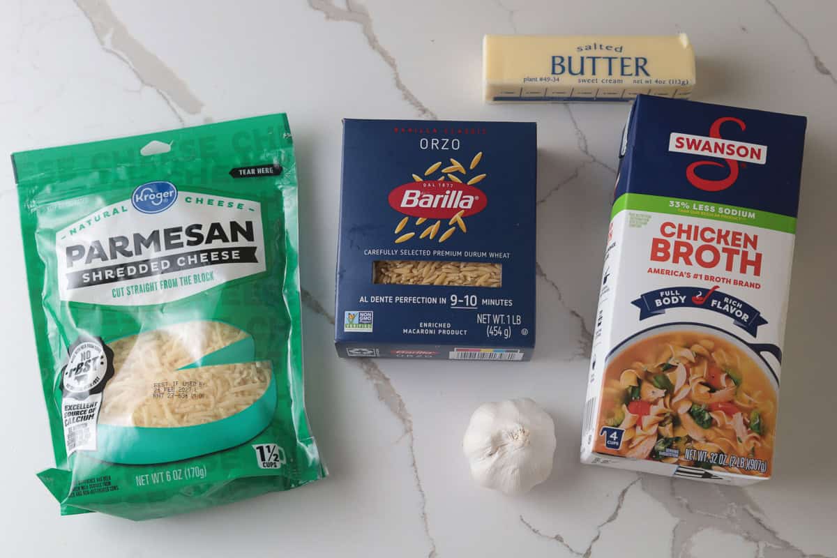 Ingredients for orzo in chicken broth, including boxed orzo pasta, chicken broth, shredded parmesan cheese, a stick of butter, and a bulb of garlic laid out on a marble countertop.