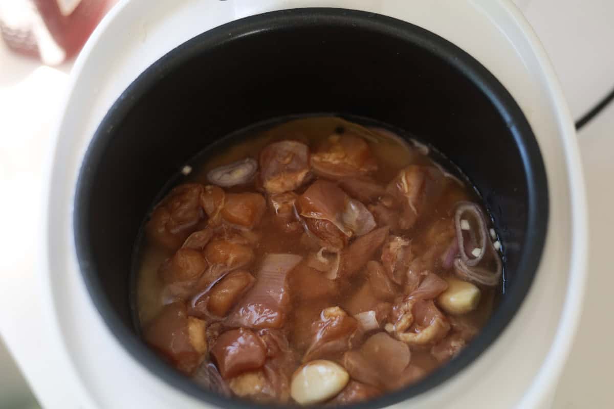 Raw cubed chicken pieces, sliced shallots, and whole garlic cloves mixed in water inside rice cooker pot.