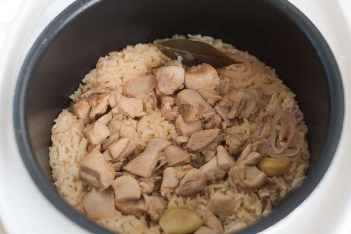 Cooked chicken adobo pieces and seasoned rice mixed together inside a rice cooker pot, garnished with a bay leaf, sliced shallots, and garlic.