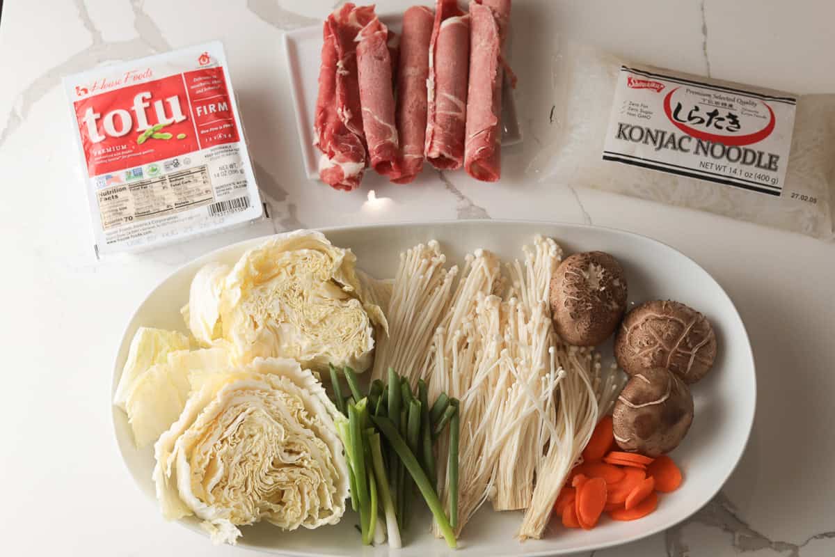 Fresh ingredients laid out on a counter, including packaged tofu, thinly sliced raw beef, konjac noodles, napa cabbage, enoki mushrooms, shiitake mushrooms, carrots, and green onions.
