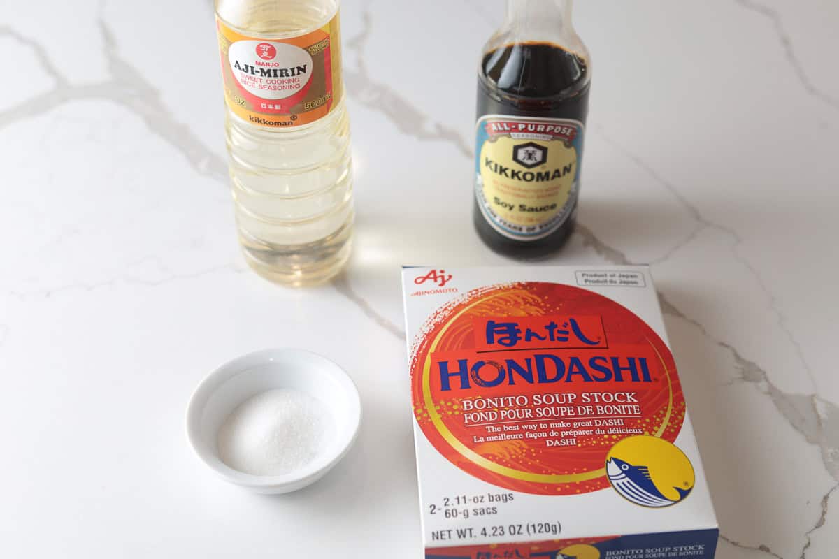 Sukiyaki seasoning ingredients arranged on a counter: a bottle of mirin, a bottle of soy sauce, a small dish of sugar, and a box of Hondashi soup stock.