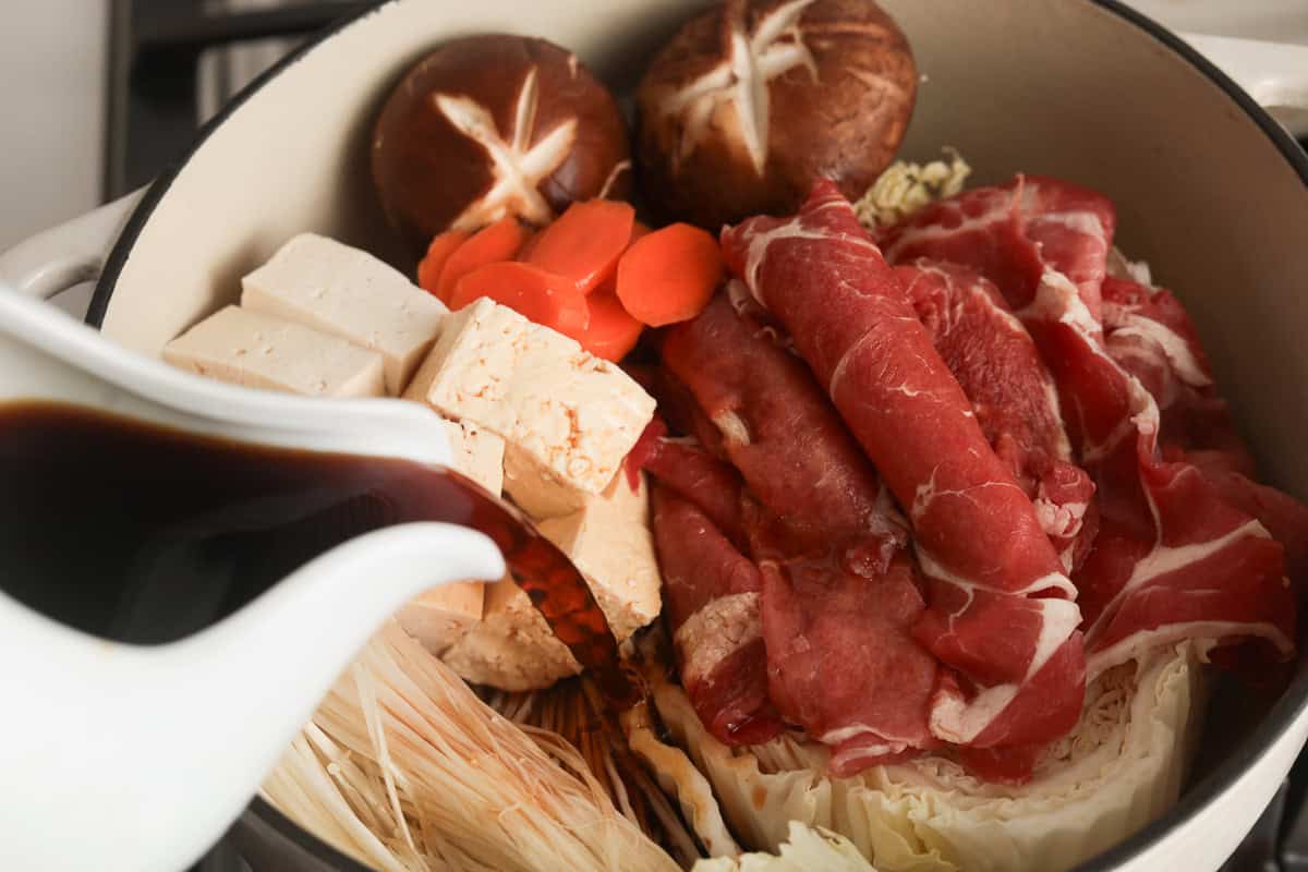 Sukiyaki sauce being poured from a white pitcher onto neatly arranged raw ingredients, including thinly sliced beef, tofu, carrots, cabbage, and mushrooms in a white pot.