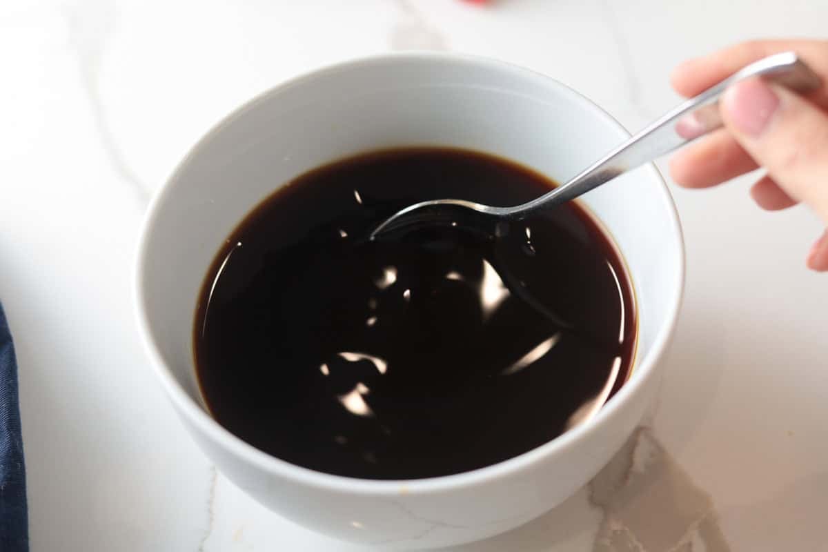 A hand stirs a dark, glossy soy-based sauce in a white ceramic bowl using a metal spoon.