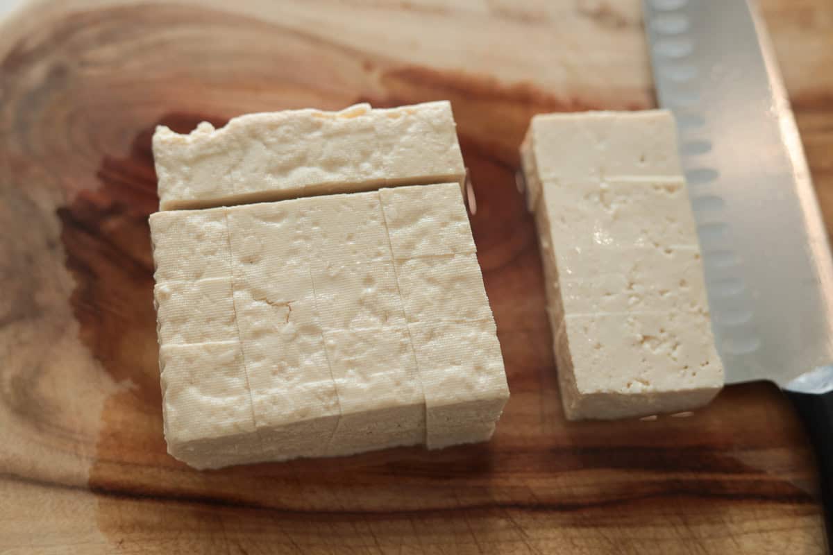 Block of firm white tofu being sliced into cubes on a wooden cutting board.