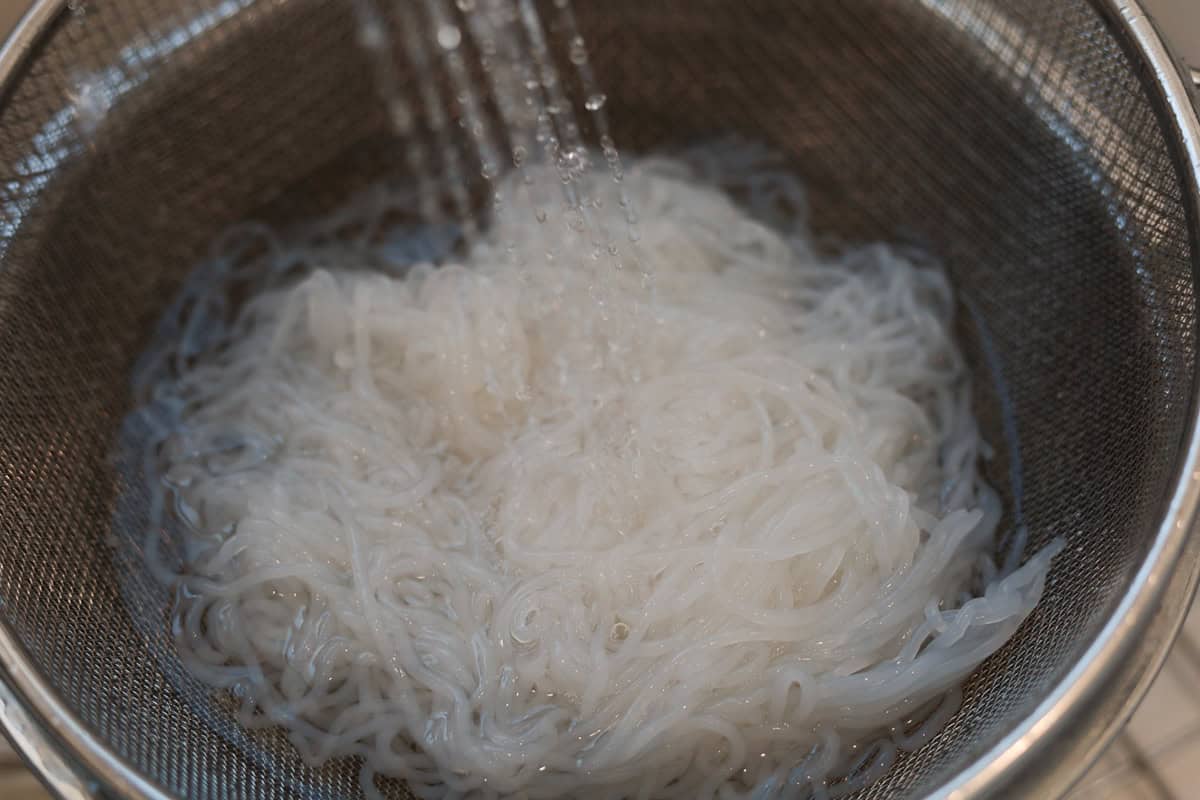 Translucent shirataki konjac noodles being rinsed under running water in a metal mesh strainer.