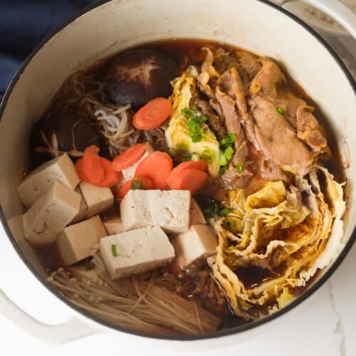 A square-cropped, top-down shot focusing tightly on a simmering pot of Japanese beef sukiyaki with mushrooms, tofu, vegetables, and sliced beef.