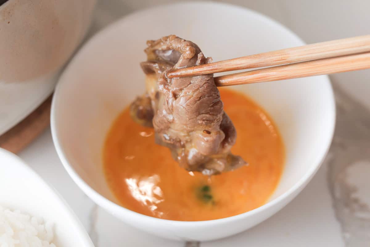 Wooden chopsticks dipping a cooked slice of beef into a small white bowl of whisked raw egg yolk.
