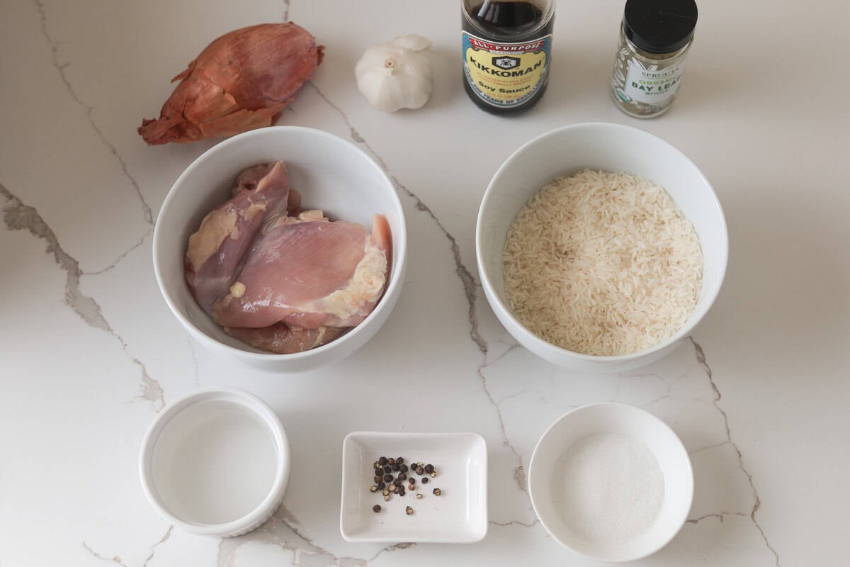 Flat lay of chicken adobo ingredients on a countertop including raw chicken thighs, white rice, a whole shallot, garlic, soy sauce, a jar of bay leaves, vinegar, peppercorns, and sugar.