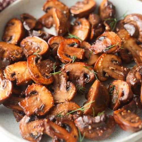 Close-up of sautéed, sliced brown mushrooms garnished with fresh green dill in a grey bowl.