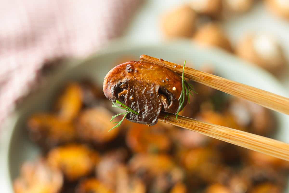 Wooden chopsticks holding up a single, glossy sautéed mushroom slice garnished with a sprig of dill.