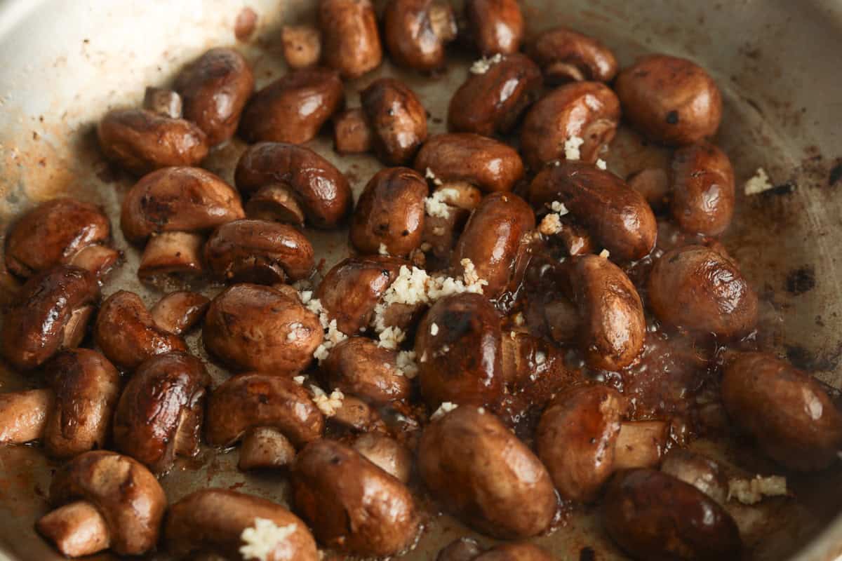 Whole brown mushrooms cooking with finely minced garlic in a stainless steel skillet.