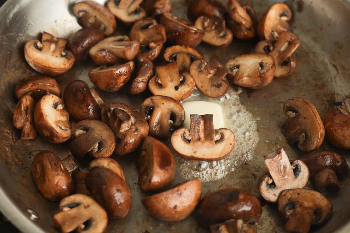 Sliced brown mushrooms sautéing in a pan around a melting pat of butter.